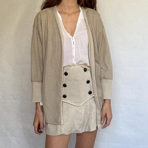 May include: A beige cardigan with long sleeves and a white blouse. The outfit includes a beige skirt with black buttons. The model is wearing blue boots. The clothing is neutral in color.