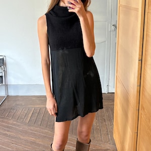 May include: A sleeveless black dress with a ribbed knit turtleneck bodice and a flowing skirt. The dress is paired with knee-high brown suede boots. The image is taken indoors with a wooden floor and a mirror.
