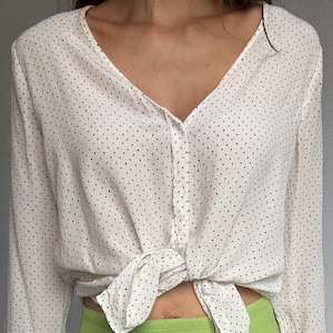 Puede incluir: Una blusa color crema de manga larga con cuello en V y pequeños lunares negros. La blusa está atada a la cintura. Combinada con una falda verde brillante.