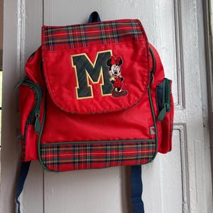 May include: A red backpack featuring a plaid pattern on the top flap and bottom panel. The front flap displays a large letter M and an embroidered Minnie Mouse character. The bag includes dark blue straps and side pockets with zipper closures.