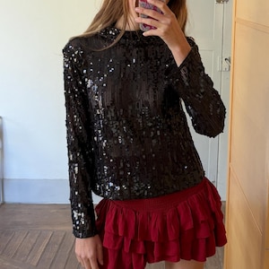 Op de afbeelding: Een zwarte top met lange mouwen en pailletten, gecombineerd met een bordeauxrode mini-rok met ruches. De top is bedekt met glanzende zwarte pailletten. De rok heeft meerdere lagen gerimpelde stof. De outfit combineert texturen en kleuren.