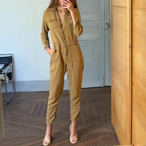 Op de afbeelding: Een bruin-beige jumpsuit met lange mouwen, een knoopsluiting en zakken. De outfit wordt gecompleteerd met witte sandalen met hakken. De foto is een spiegelbeeld.