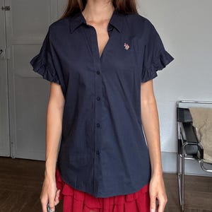 Può includere: Una camicia blu navy con bottoni e maniche corte a balze. La camicia presenta un piccolo logo ricamato sul petto. La modella indossa una gonna rossa. Lo sfondo è una stanza dai toni neutri con una porta e una sedia.