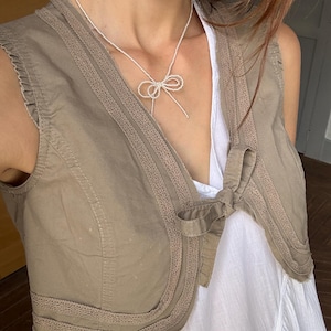Puede incluir: Un chaleco color canela con costuras decorativas y cierre de lazo sobre un vestido blanco. Se lleva un delicado collar de cuentas blancas con forma de lazo. El atuendo es informal y elegante.