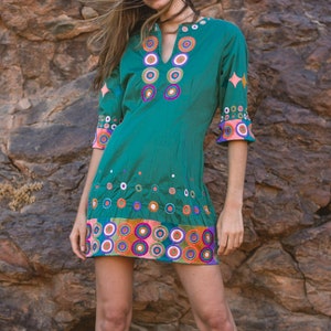 May include: A teal-green tunic dress with colourful embroidered circles and starbursts. The dress has a V-neck and three-quarter sleeves with pink trim. The hemline and sleeves are adorned with vibrant, multi-coloured circles. The person is wearing brown suede boots.