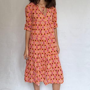 Könnte beinhalten: Midikleid mit geometrischem Muster in Orange, Rosa und Braun. Das Kleid hat einen V-Ausschnitt mit Bindeband, kurze Ärmel und einen ausgestellten Rock. Das Muster wiederholt sich auf dem gesamten Kleid.