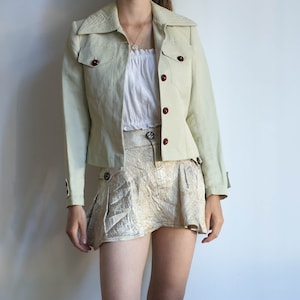 Pode incluir: Casaco verde claro com botões vermelhos escuros e minissaia a condizer. O casaco tem gola e dois bolsos frontais. A saia tem um design plissado e um padrão texturizado dourado. Uma blusa branca completa o conjunto.