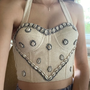 May include: Cream-colored halter top with seashell embellishments. The top features a sweetheart neckline and is adorned with small, white seashells outlined with black beads. The design is repeated in a pattern across the bodice.