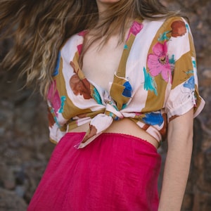 May include: A floral print top with a knotted front and short sleeves paired with bright pink pants. The top features a pattern of pink, blue, and brown flowers on a cream background. The pants are a vibrant, solid pink color.