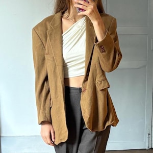May include: A brown blazer with a single button closure, paired with dark gray trousers. The outfit includes a white top and light-colored sandals. The blazer has a classic design with a notched lapel.