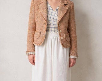 Könnte beinhalten: Ein hellbrauner Tweed-Blazer mit Reverskragen, zwei Vordertaschen und Knopfverschlüssen. Der Blazer wird mit einem karierten Oberteil und einer weißen Hose mit weitem Bein kombiniert. Das Outfit ist ein klassischer, eleganter Stil.