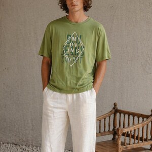 Könnte beinhalten: Eine Person trägt ein grünes T-Shirt mit einem rautenförmigen Aufdruck und dem Text "MOVING TOGETHER CONCEPT", kombiniert mit einer weißen Leinenhose und schwarzen Schuhen. Ein Holzstuhl befindet sich im Hintergrund.