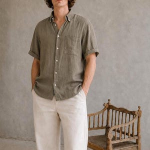May include: A person wearing a short-sleeved olive green button-down shirt and cream-colored pants. The person is wearing black leather shoes. An antique wooden chair is in the background.
