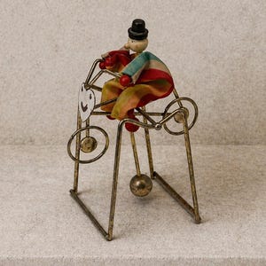 May include: A vintage toy featuring a small figure in a colorful outfit and a black top hat, seated on a brass-colored metal frame. The toy has a whimsical design with a smiling face on the side and a small ball beneath the seat.