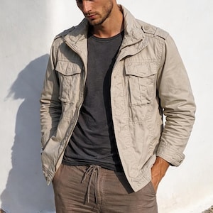Puede incluir: Chaqueta beige de estilo militar con múltiples bolsillos, usada sobre una camiseta gris oscuro y combinada con pantalones marrones con cordón. El atuendo es casual y elegante, adecuado para el uso diario.