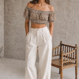 May include: An off-the-shoulder crop top with a leopard print pattern is paired with high-waisted, wide-leg white pants. Black shoes complete the outfit. A wooden chair with a woven seat is in the background.