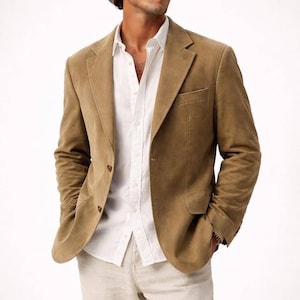 May include: A tan corduroy blazer with a notched lapel, worn over a white button-down shirt and light-colored pants. The blazer has a two-button closure and a chest pocket. The outfit is a classic, casual style.