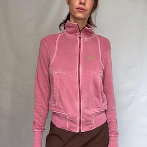 K&ouml;nnte beinhalten: Rosa Jacke mit Rei&szlig;verschluss, hohem Kragen und zwei Vordertaschen. Die Jacke hat ein verwaschenes Aussehen und ein kleines Logo auf der Brust. Das Model tr&auml;gt einen braunen Rock.
