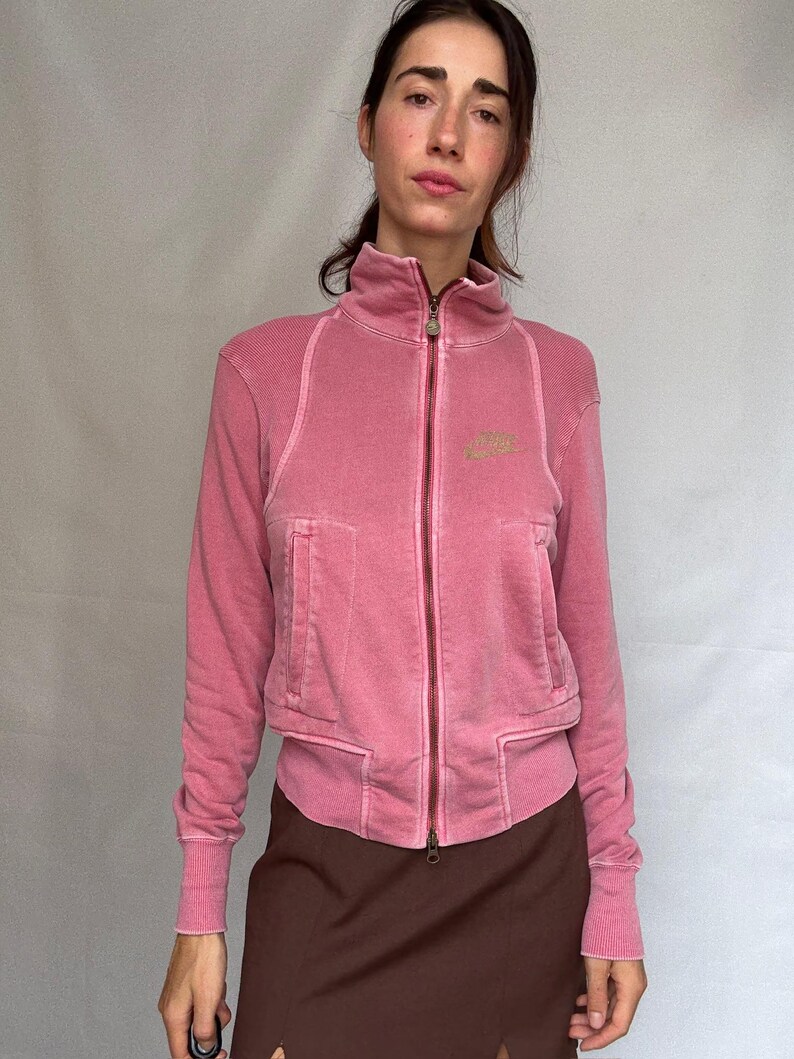 K&ouml;nnte beinhalten: Rosa Jacke mit Rei&szlig;verschluss, hohem Kragen und zwei Vordertaschen. Die Jacke hat ein verwaschenes Aussehen und ein kleines Logo auf der Brust. Das Model tr&auml;gt einen braunen Rock.