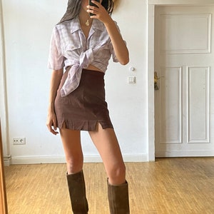 May include: A woman wearing a light purple plaid shirt tied at the waist, a brown mini skirt with side slits, and brown knee-high boots.