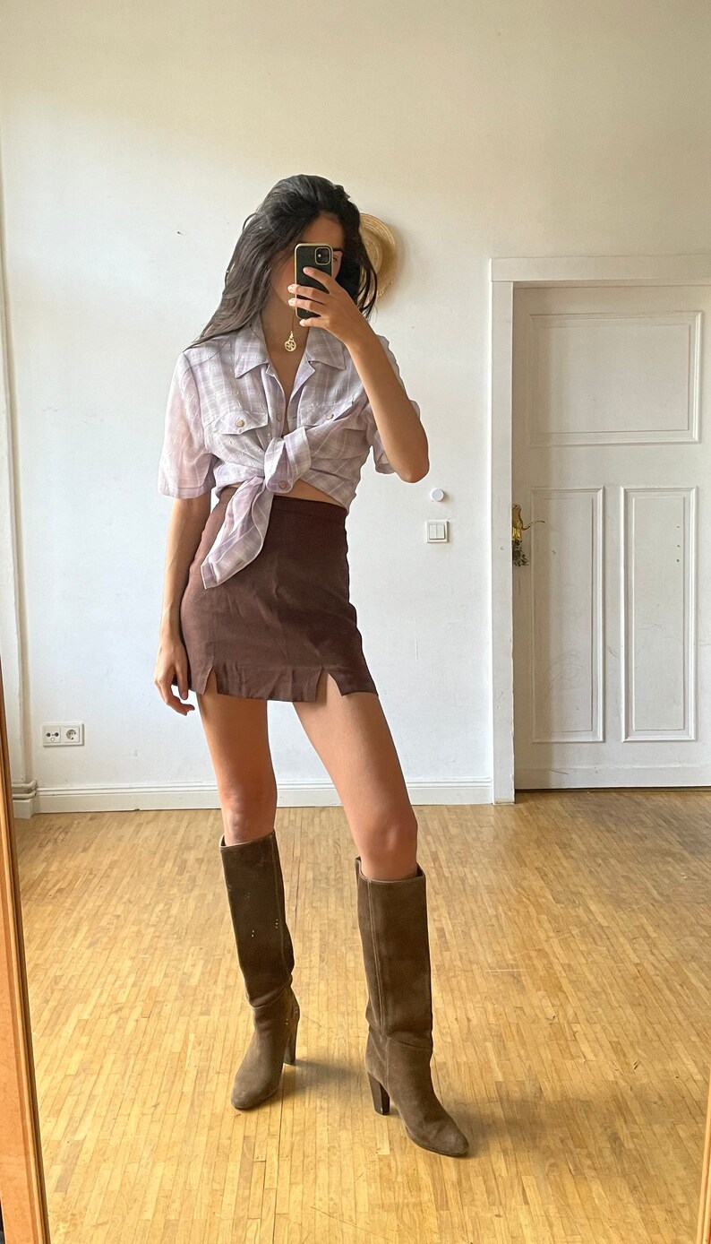 May include: A woman wearing a light purple plaid shirt tied at the waist, a brown mini skirt with side slits, and brown knee-high boots.