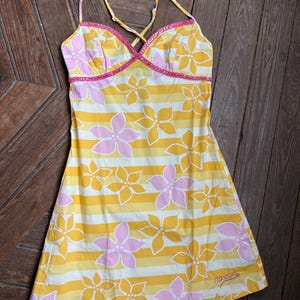May include: A yellow, white, and pink floral print dress with thin straps. The dress features a striped pattern with large flower designs. The neckline and straps are trimmed with pink detailing. The brand name "ripcurl" is printed on the bottom.