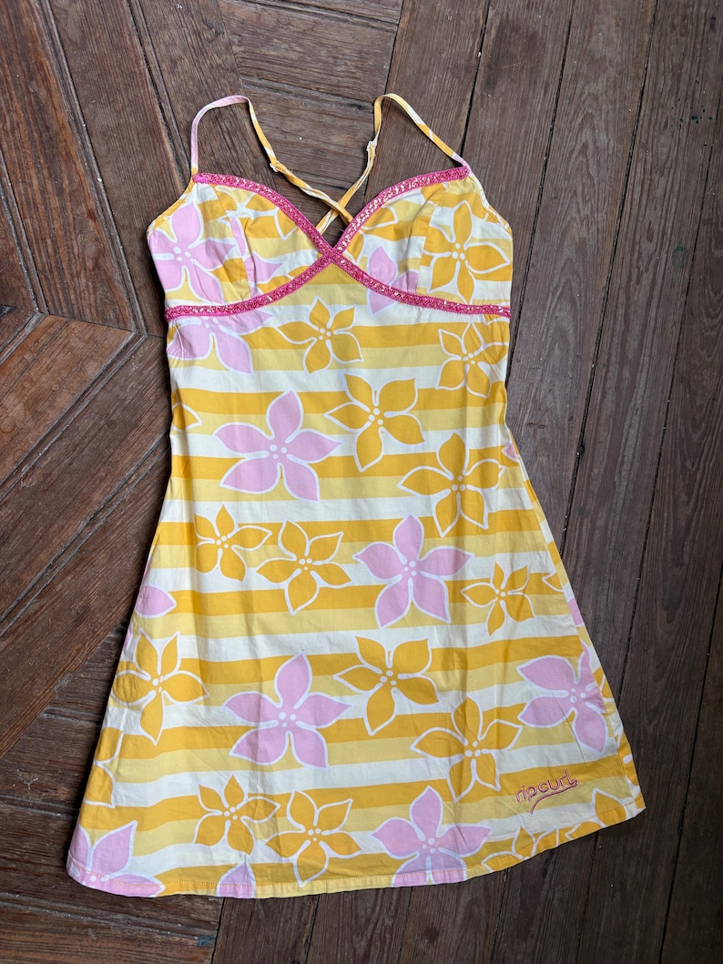 May include: A yellow, white, and pink floral print dress with thin straps. The dress features a striped pattern with large flower designs. The neckline and straps are trimmed with pink detailing. The brand name "ripcurl" is printed on the bottom.