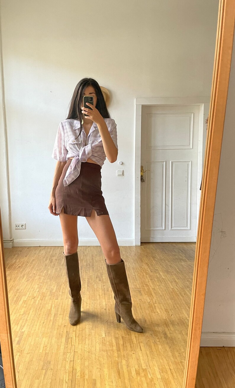 May include: A woman wearing a light purple and white plaid shirt tied at the waist, a brown mini skirt, and brown knee-high boots. She is standing in front of a mirror in a room with wood floors.