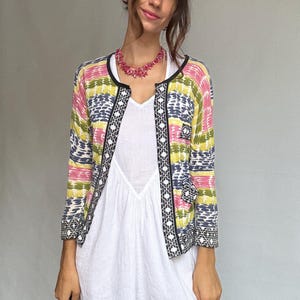 May include: A colorful, patterned cardigan with black trim and a white dress. The cardigan features pink, yellow, and blue horizontal stripes. The dress is a simple white design. A pink necklace is also visible.