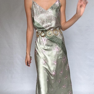 May include: A light green slip dress with a V-neck and spaghetti straps. The dress features a floral pattern with pink flowers and a gold-coloured belt. The dress is made of a silky material.
