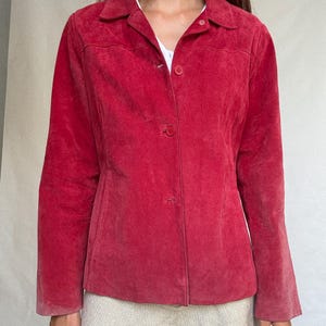 May include: A red suede jacket with a collared neckline and button-up closure. The jacket has long sleeves and a tailored fit. The color is a vibrant, solid red, and the material appears soft and textured. A white shirt is visible underneath.