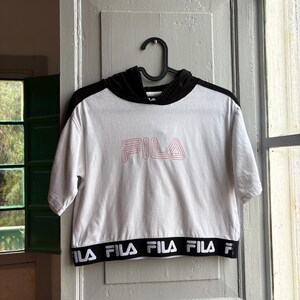 May include: White cropped hoodie with black sleeves and hood. The word "FILA" is printed in pink on the front. The bottom band features the brand name in black and white. The shirt is hanging on a black hanger.