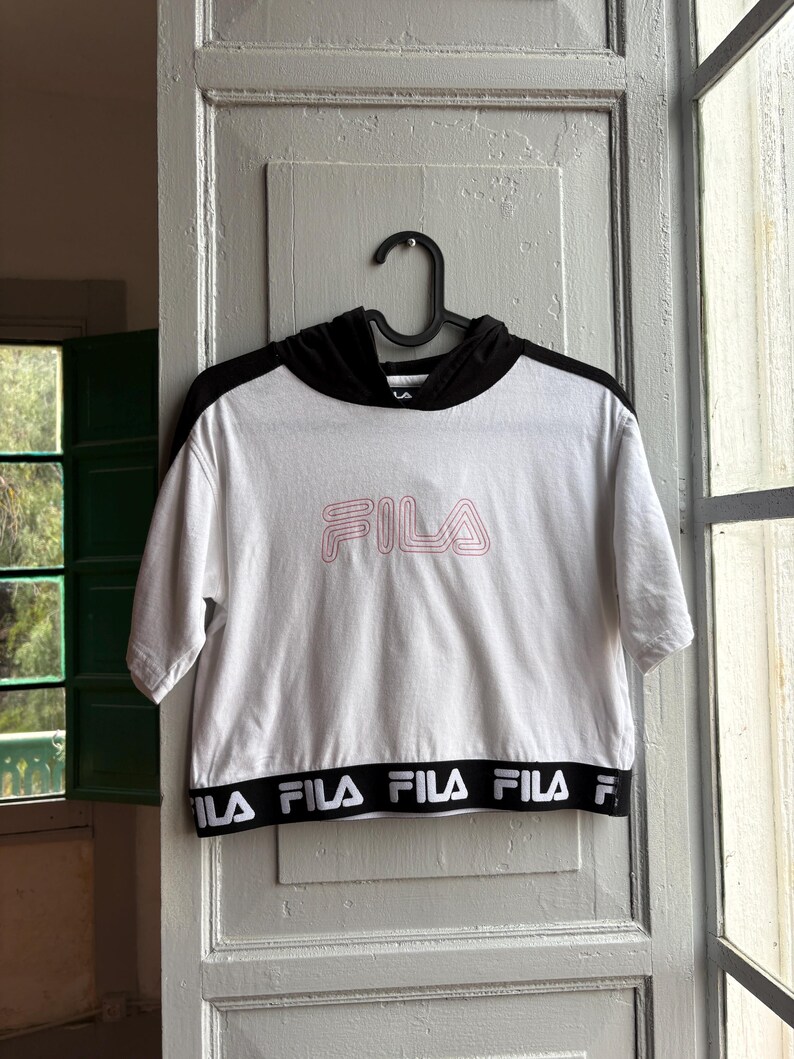 May include: White cropped hoodie with black sleeves and hood. The word "FILA" is printed in pink on the front. The bottom band features the brand name in black and white. The shirt is hanging on a black hanger.