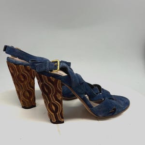 May include: Blue suede high-heeled sandals with a crisscross strap design. The heels are a dark brown with a carved pattern. The sandals have a buckle closure with gold-colored hardware.