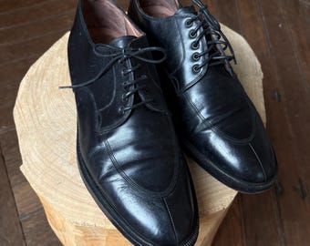 Puede incluir: Un par de zapatos de vestir de cuero negro con cordones. Los zapatos se exhiben sobre una superficie de madera de color marrón claro. Tienen un diseño clásico con punta redonda y tacón bajo.