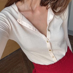 Peut inclure: Un cardigan rose clair boutonné avec des boutons dorés. Il est associé à une jupe rouge avec une taille smockée. L'ensemble est posé sur un sol en bois.