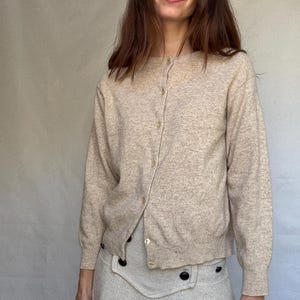 May include: A beige cardigan with gold buttons. The long-sleeved sweater is buttoned up. The person is wearing a matching beige skirt with black buttons.