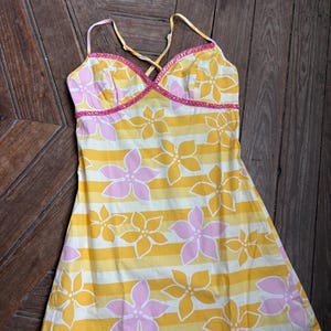 May include: A yellow and white striped dress with pink and yellow floral patterns. The dress has thin spaghetti straps and a pink trim along the neckline. The Rip Curl logo is visible near the hem.