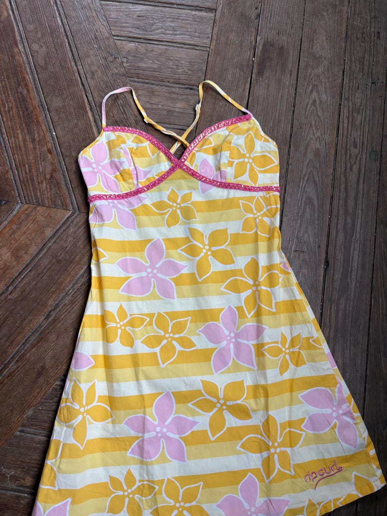 May include: A yellow and white striped dress with pink and yellow floral patterns. The dress has thin spaghetti straps and a pink trim along the neckline. The Rip Curl logo is visible near the hem.
