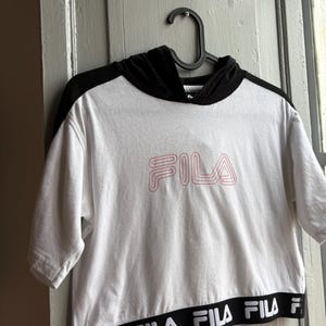 May include: White crop top with a black hood and short sleeves. The word "FILA" is printed in pink on the front. The bottom hem features a black band with the brand name in white. The top is hanging on a black hanger.