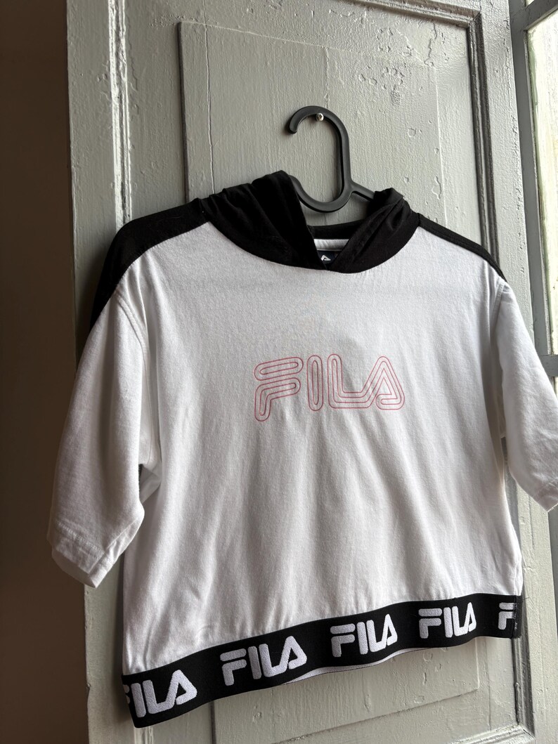 May include: White crop top with a black hood and short sleeves. The word "FILA" is printed in pink on the front. The bottom hem features a black band with the brand name in white. The top is hanging on a black hanger.