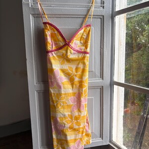 May include: A yellow sundress with pink and white floral and striped patterns hangs on a hanger. The dress has thin straps and a pink trim along the neckline. The dress is hanging in front of a window.