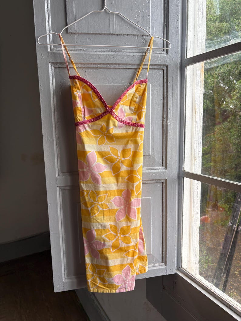 May include: A yellow sundress with pink and white floral and striped patterns hangs on a hanger. The dress has thin straps and a pink trim along the neckline. The dress is hanging in front of a window.