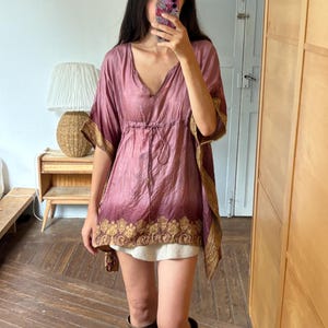 Può includere: Una tunica color malva con scollo a V e coulisse in vita. Il capo presenta maniche corte e ampie e un motivo paisley color oro lungo l'orlo e i bordi delle maniche. Il tessuto ha una lucentezza sottile e una consistenza leggermente stropicciata.