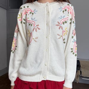 May include: Cream-colored cardigan sweater with floral embroidery in pink, yellow, blue, and green. The button-down sweater has long sleeves and a round neckline. The bottom of the sweater is paired with a red ruffled skirt.