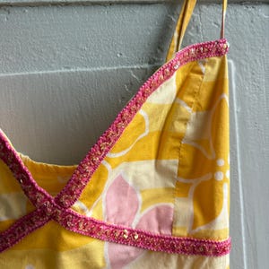 May include: A yellow sundress with a floral pattern in white and pink. The dress has thin straps and is trimmed with pink and gold sequined trim. The fabric is a light cotton.