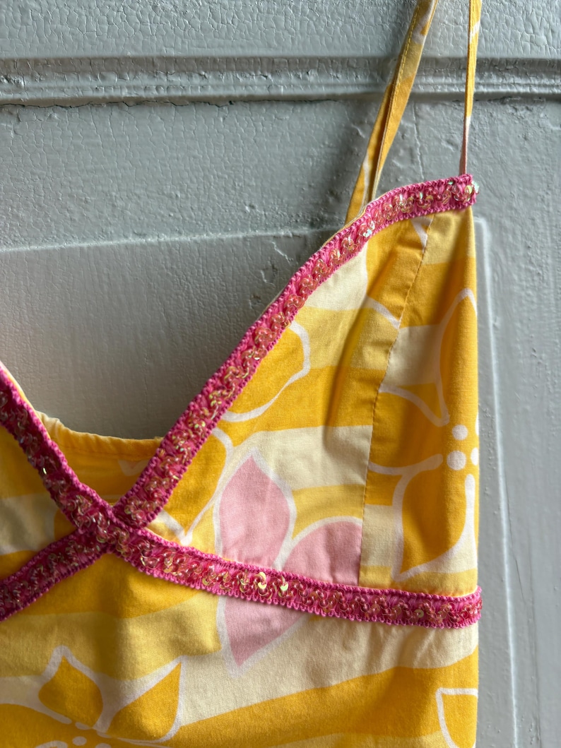 May include: A yellow sundress with a floral pattern in white and pink. The dress has thin straps and is trimmed with pink and gold sequined trim. The fabric is a light cotton.