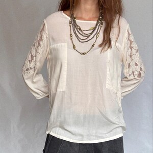 May include: Cream-colored blouse with lace sleeves and two front pockets. The blouse is paired with a dark gray skirt. A multi-strand necklace with leaf and bead accents is worn.