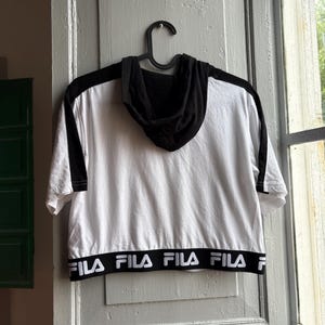 May include: White and black hooded crop top with short sleeves. The top features black accents on the shoulders and hood, with the brand name "FILA" printed in white on the bottom hem. The top is hanging on a black hanger.