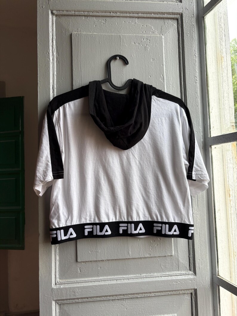 May include: White and black hooded crop top with short sleeves. The top features black accents on the shoulders and hood, with the brand name "FILA" printed in white on the bottom hem. The top is hanging on a black hanger.